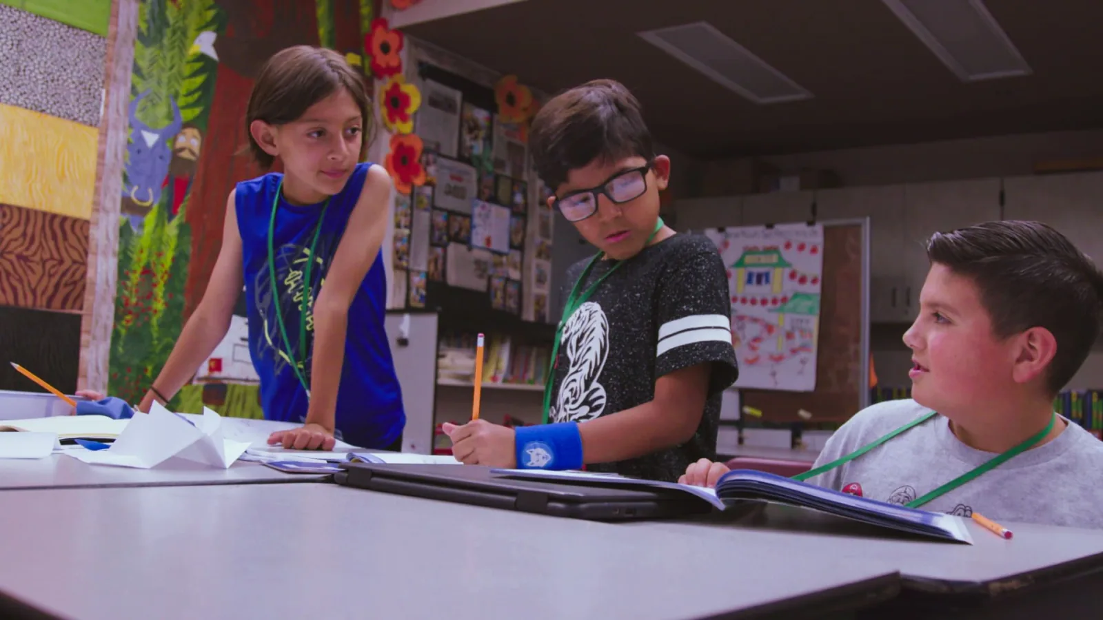 Three students stand and talk through their notebooks together at a classroom table